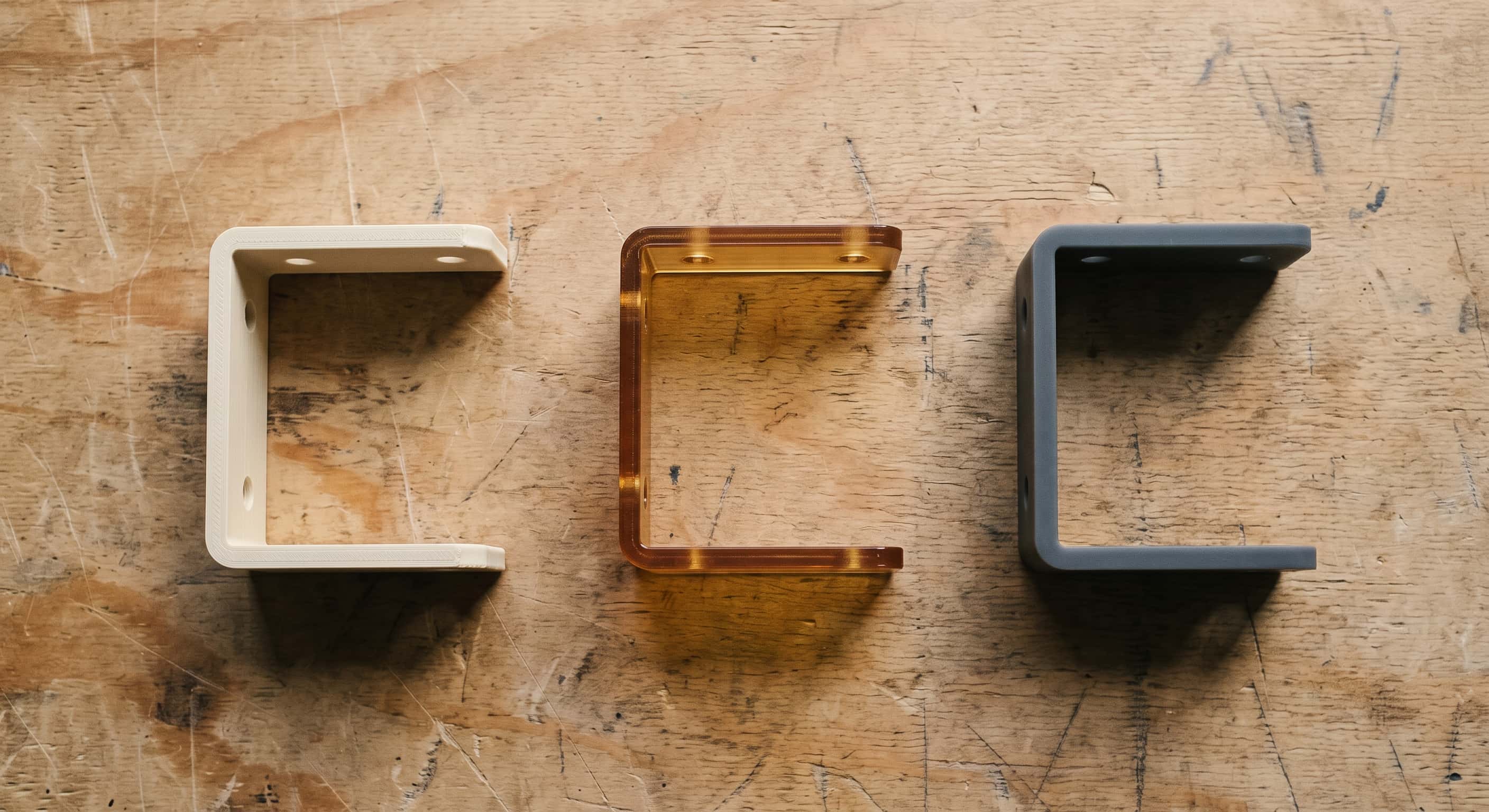 Three identical L-shaped brackets photographed from above on a scuffed plywood bench, printed in cream PLA, translucent amber PETG and dark slate resin
