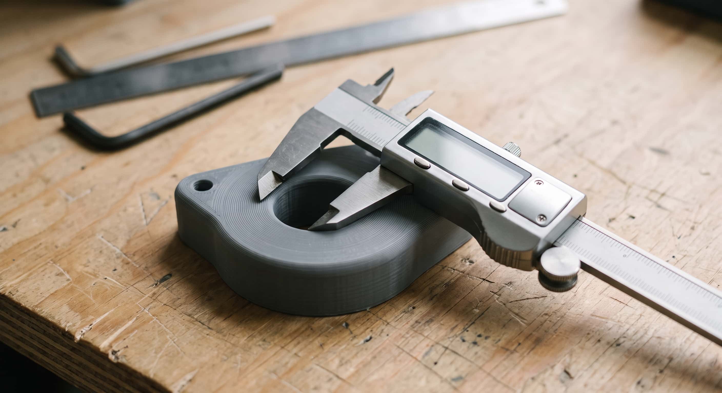 Digital calipers with the jaws closed across the bore of a grey printed part on a plywood bench, hex keys and a steel rule out of focus behind